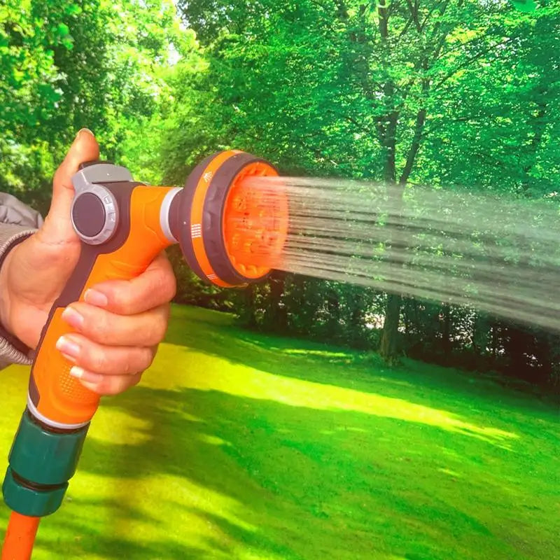 Hand holding an orange garden hose spray nozzle with water streaming out in a green garden setting.