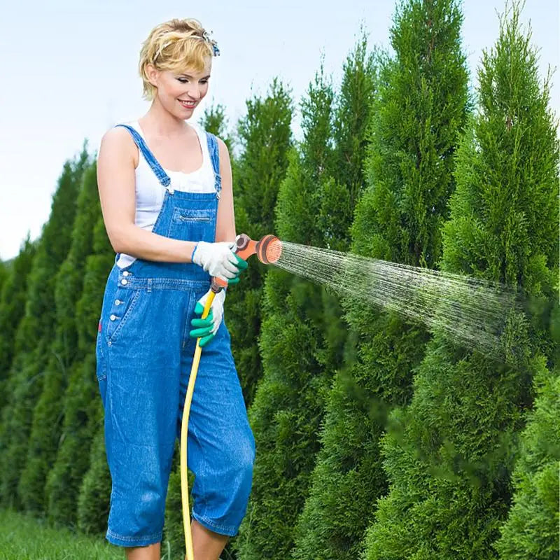 Woman using garden hose spray nozzle for watering tall bushes in garden.